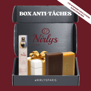 Box Anti-Tâches Nirlys Paris présentée dans une boîte noire ouverte, contenant un sérum éclat & anti-tâche, deux savons (curcuma et exfoliant), et un coffret cadeau blanc noué d’un ruban doré. Une mention “Une surprise vous attend dans votre box” figure en haut à droite.